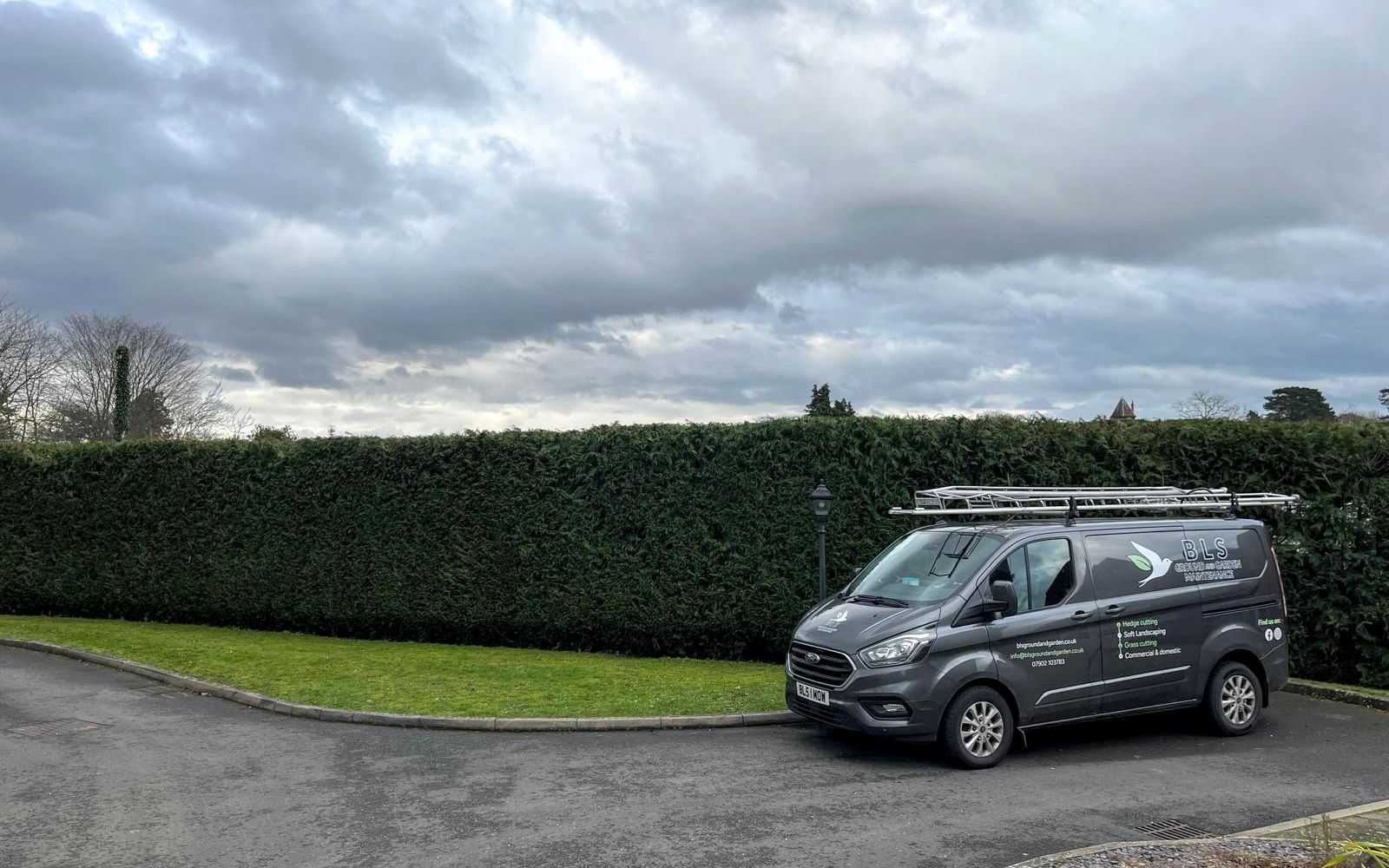 A BLS Ground & Garden van parked beside a long, neatly trimmed hedge.